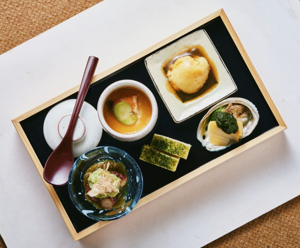 Assorted Japanese appetizers including chawanmushi, tofu, and seasonal dishes in a wooden tray