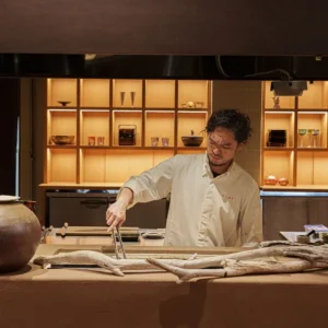 Chef preparing a refined dish in a modern Japanese kitchen with illuminated wooden shelves at a luxury ryokan with private onsen on Awaji Island.