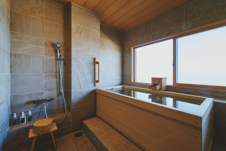 Japanese‑style deluxe room bathroom with cypress soaking tub, stone tiles, and soft natural light at a luxury ryokan with private onsen on Awaji Island.